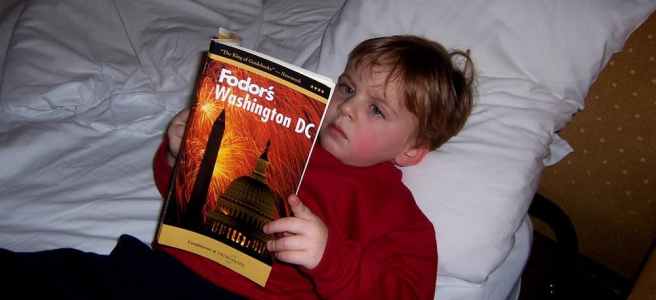 A child lies on a bed reading a Fodor's guidebook to Washington, DC.